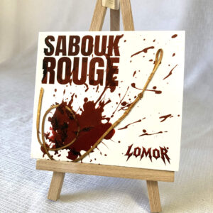 The front CD cover is exposed on an easel with its big blood stain in the bottom left. The Lomor logo is on the bottom right whereas the title "Sabouk Rouge" seats on the upper left.